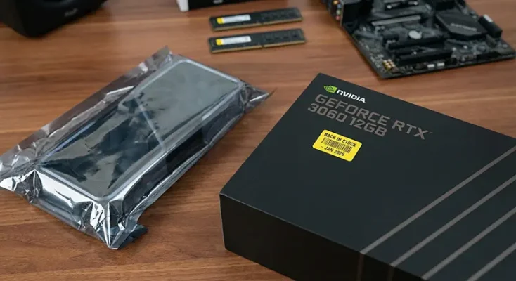 Product box of the NVIDIA GeForce RTX 3060 12GB graphics card sitting on a wooden desk next to a PC case.